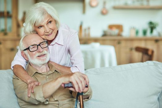 Happy elderly couple having rest in living room while woman embracing his husband. Man holding walking stick. Family and relationships concept