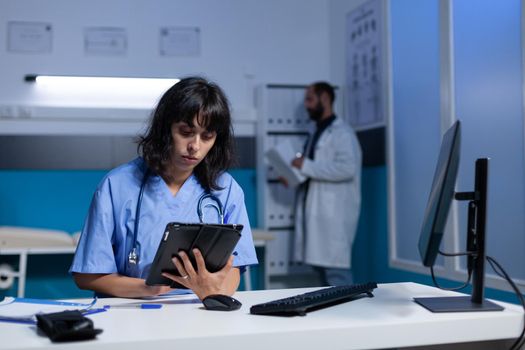 Woman working as nurse with digital tablet for checkup at night. Medical assistant using modern device and computer on desk for healthcare and treatment, working late. Person with job