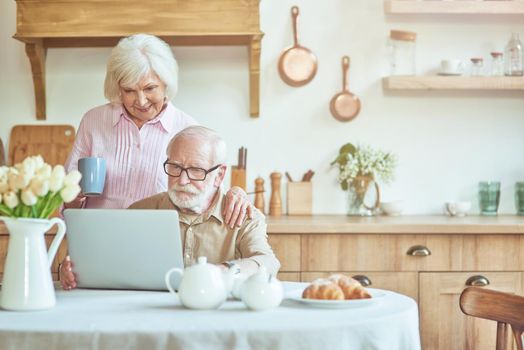 Happy senior wife standing near her husband while looking at laptop screen at the kitchen. Domestic lifestyle concept