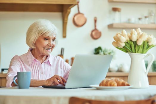 Happy senior female pensioner using computer while sitting on the kitchen in the morning while typing. Domestic lifestyle concept