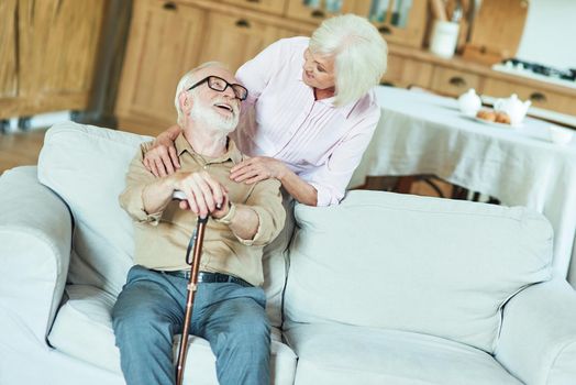 Happy senior man with walking stick sitting on sofa and looking at his wife at their home. Family and relationships concept