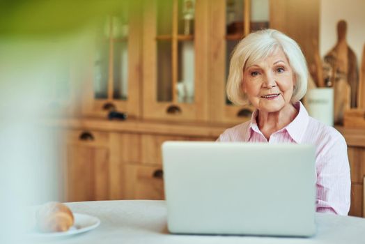 Waist up of smiling senior woman sitting in the room at the table while working with laptop. Domestic lifestyle concept