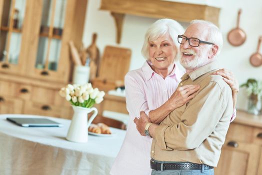 Waist up of happy senior man and woman hugging while posing at camera at their kitchen. Lifestyle concept