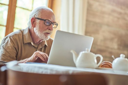 Male retiree in glasses carefully looking at laptop screen while working at home. Lifestyle concept