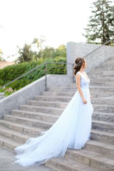 Back view of european bride walking on concrete stairs and wearing white dress. Concept of bridal fashion and wedding vogue.