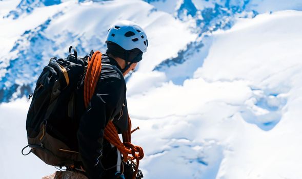 A young man traveler is engaged in mountaineering. Hiker in a helmet, with a rope climbs to the top, against the backdrop of a stunning view with snow-capped mountains.