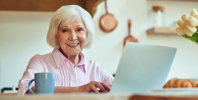 Waist up of smiling elderly lady typing on laptop while sitting at the table at home. Domestic lifestyle concept