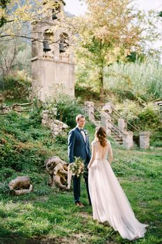 Bride goes to groom with a bouquet near the old chapel in the garden. High quality photo