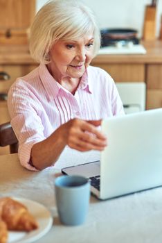 Smiling elderly female using laptop while looking at screen at the kitchen at home. Domestic lifestyle concept