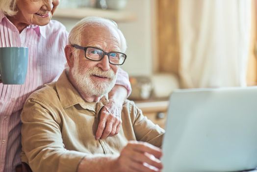 Happy elderly man in glasses working on laptop with her wife with cup standing near him at the kitchen. Domestic lifestyle concept