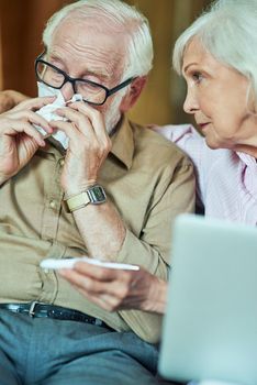 Close up of senior wife taking care of her spouse while holding thermometer and sitting next to him. Care and health concept