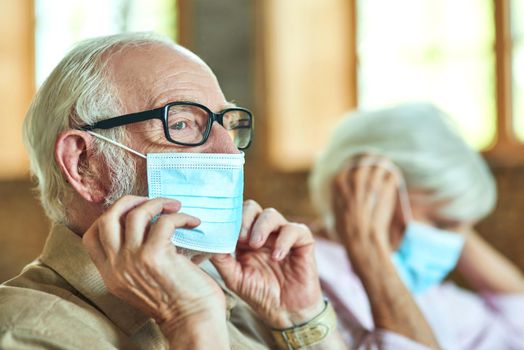 Gray-haired elderly man with glasses adjusting protective mask with her wife sitting on the background at home. Care and health concept