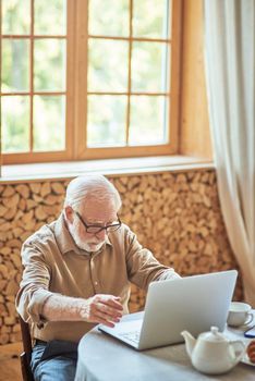 Elderly man in glasses sitting at the table while working at laptop in his cozy home. Lifestyle concept