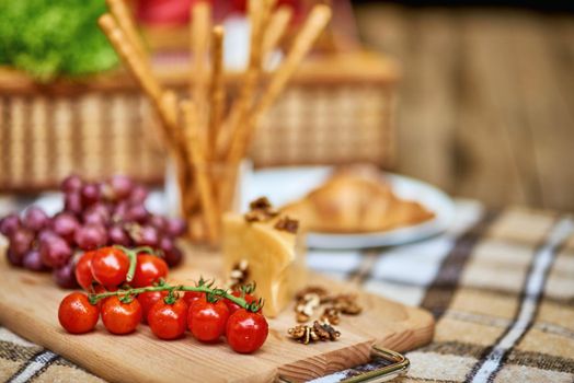 Tomatoes, grapes and cheese on a cutting wooden board on checkered plaid with a wicker basket on the background. Rest and vacation lifestyle concept