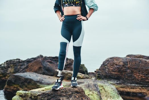 Cropped photo of disabled woman in sports clothing with prosthetic leg standing on the stone and keeping arms on her hips. Disabled Sportsman. Healthy lifestyle. Happiness concept