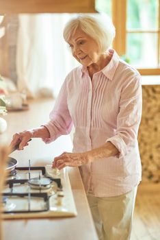 Smiling senior woman turning on the stove and setting the frying pan to make breakfast at home. Domestic lifestyle concept