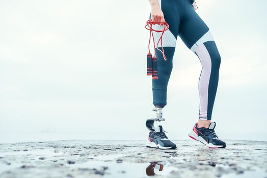 Doing my best. Cropped image of disabled woman in sports clothing with prosthetic leg holding skipping rope while standing in front of the sea. Disabled Sportsman. Healthy lifestyle. Cardio