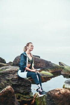 Enjoying nature and music. Happy disabled athlete woman in sportswear with prosthetic leg sitting on the stone and listening music. Sport concept. Music concept. Disabled Sportsman.