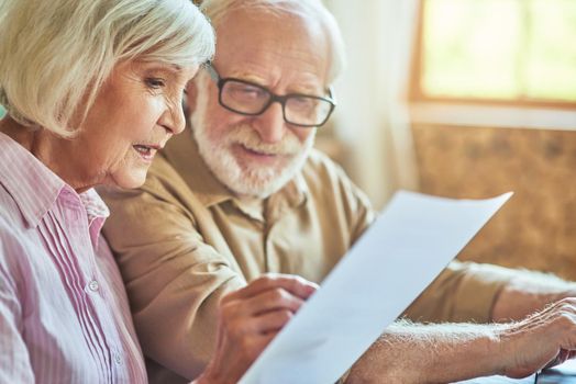 Close up of smiling middle aged couple holding and reading good news in document at home. Family and household concept