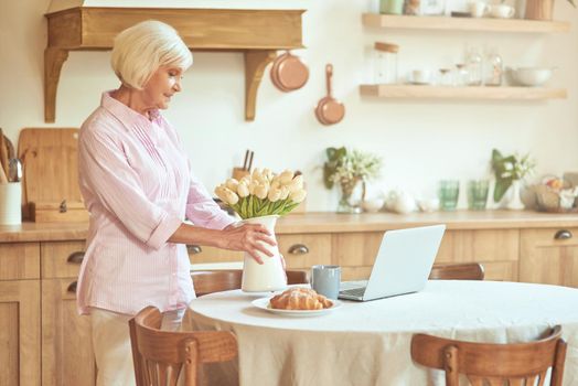 Happy elderly woman putting a vase of flowers on the table in the kitchen in the morning. Domestic lifestyle concept