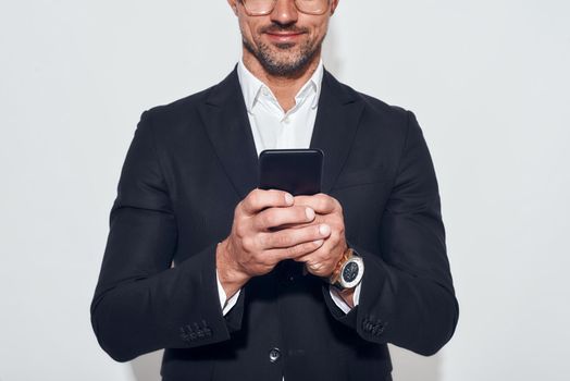 Great news. Portrait of handsome and cheerful bearded man in classic wear and eyeglasses holding smart phone and looking at it while standing against grey background. Digital concept. Business concept
