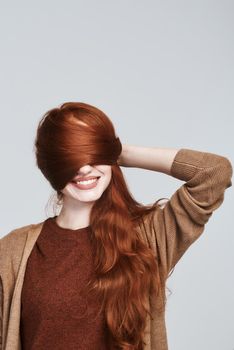 Studio shot of cheerful and young redhead woman playing with her hair and smiling while standing against grey background. Beauty concept. Happiness