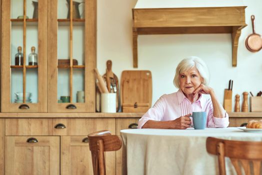 Smiling stylish gray haired lady enjoying morning coffee while holding mug at home. Domestic lifestyle concept