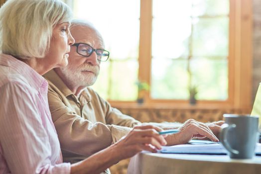 Side view of senior couple using laptop while checking bills together at home. Copy space. Family and household concept