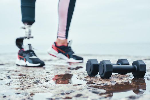 Strong and healthy. Cropped image of woman with prosthetic leg and dumbbells lying in front of the sea. Disabled Sportsman. Healthy lifestyle. Workout
