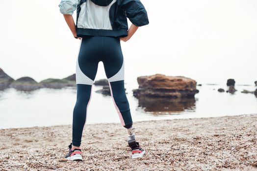 Make everyday better Cropped image of sporty and confident disabled woman in sportswear with prosthetic leg standing on the beach and keeping arms on her hips. Motivation concept. Disability concept. Disabled Sportsman