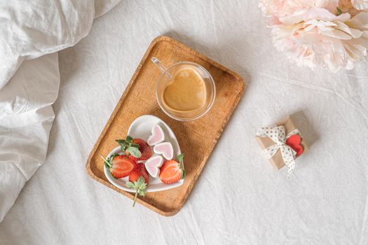 Breakfast for Valentine's Day. Heart shaped white plate with fresh strawberries, cup of coffee, gift and flowers with gift in bed. Still life composition.