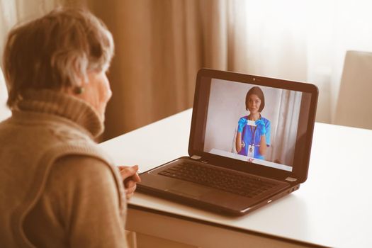 An elderly patient, retired person participates in a digital conversation with a nurse via a webcam. Aged woman talking using a video link with a female doctor, has an online consultation at home.