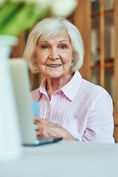 Happy senior woman enjoying morning coffee while sitting at home and using laptop. Domestic lifestyle concept