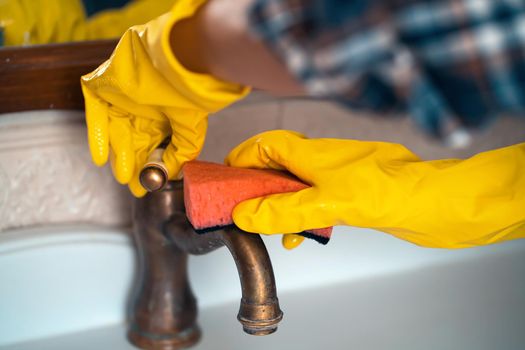 A woman in rubber gloves is cleaning the house, creating cleanliness and disinfection, wiping the faucet and wash basin in the bathroom with a sponge.