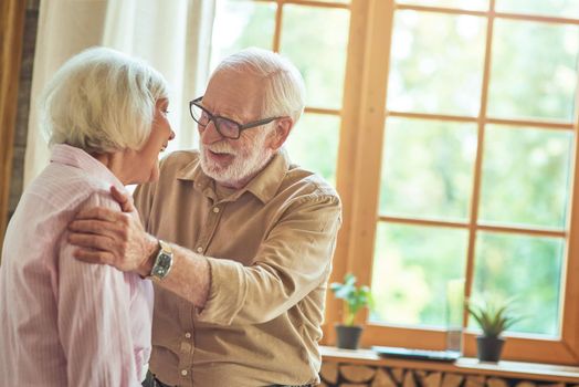 Happy elderly man hugging his wife while standing near window at home. Family and relationships concept. Copy space