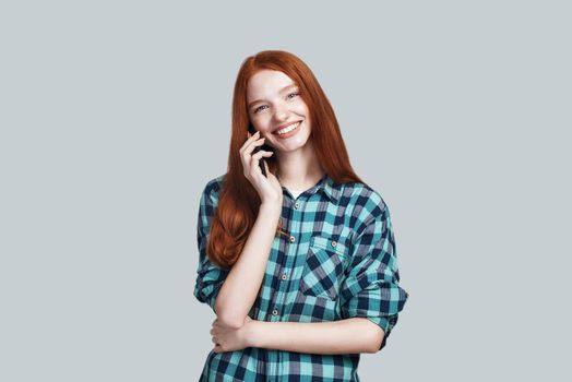 Studio shot of young and cute redhead woman in casual wear is talking on the phone and smiling while standing against grey background. Digital concept. Web banner