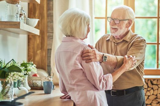 Happy senior man holding his wife by the shoulders with a window on the background in their house. Family and relationships concept