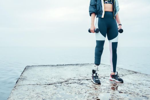 Exercising with dumbbells. Cropped image of disabled woman in sports clothing with prosthetic leg holding dumbbells while standing in front of the sea. Disabled Sportsman. Healthy lifestyle. Workout