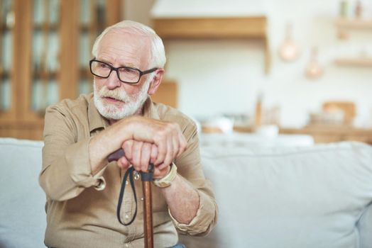 Waist up of pensive gray-haired senior man holding walking stick and looking away at home. Care and health concept. Copy space