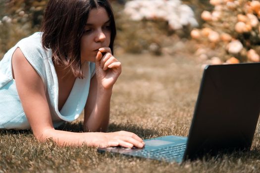 A young girl in a white dress is working on a laptop, texting on the Internet on her device, freelancing, studying, resting on a meadow in a park, lying on the grass.