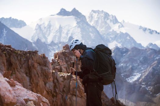 A young man traveler is engaged in mountaineering. In a helmet and with trekking poles, tourist climbs to the top, against the backdrop of a stunning view from the snow-capped mountains and rocks.