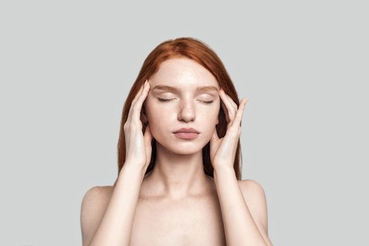 Studio shot of beautiful redhead woman keeping eyes closed and touching head with hands while standing against grey background