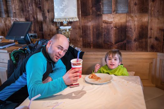 young happy father and his little son eating a pizza after a hard snowboarding day during winter vacation at beautiful ski resort
