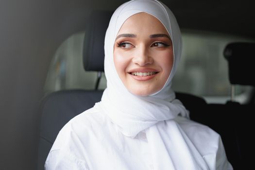 Beautiful muslim woman in white hijab sitting on the back seat of a luxury car, portrait