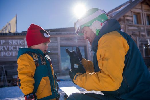 young happy father preparing his little son for the first time on a snowboard during sunny winter day at beautiful  ski resort