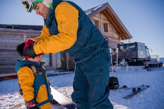 young happy father preparing his little son for the first time on a snowboard during sunny winter day at beautiful  ski resort