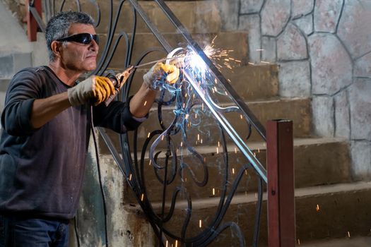 Man wearing protective glasses welds metal with welding machine in a private house. Sparks from welding fly to sides.