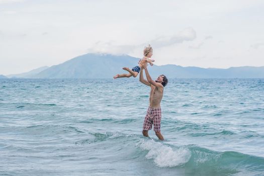 young happy father holding up in his arms little son putting him up at the beach waves wet sand having fun with the kid in Summer sunset coast.