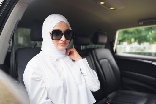 Beautiful muslim woman in white hijab sitting on the back seat of a luxury car, portrait