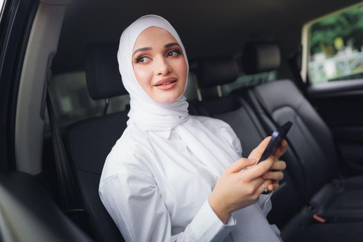 Beautiful muslim woman wearing white hijab sitting on the back seat of a car and using smartphone, close up